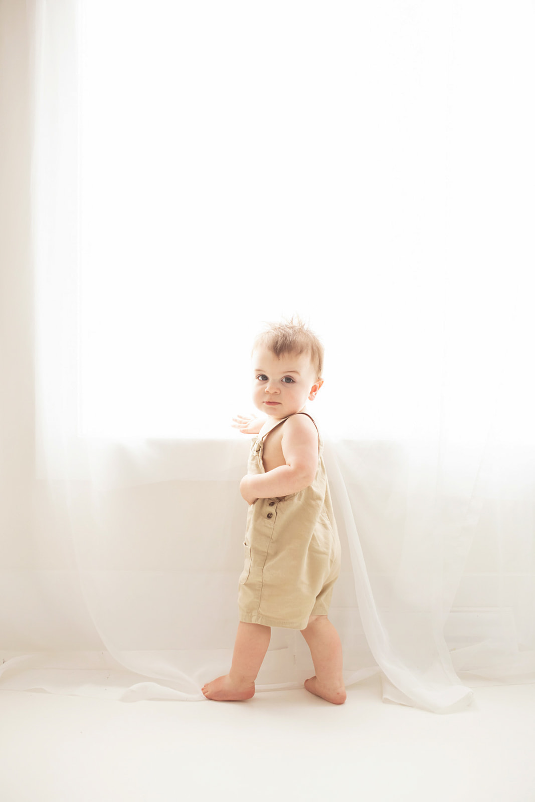 A curious toddler boy in beige overalls explores a window in a studio before visiting Pittsburgh splash pads