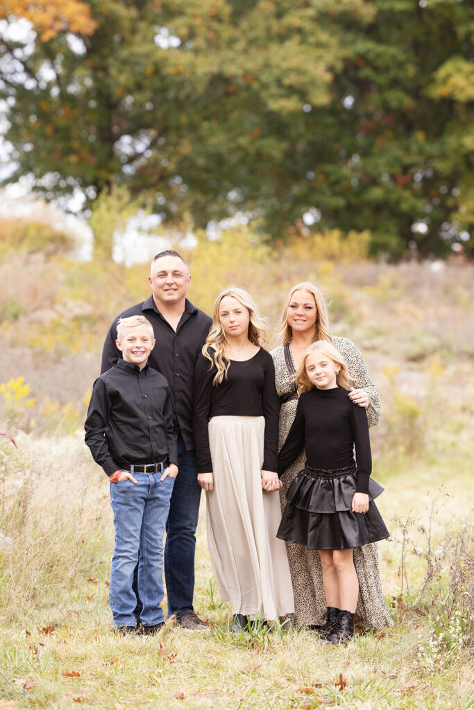 A happy family of 5 in black tops stand in a park field at sunset