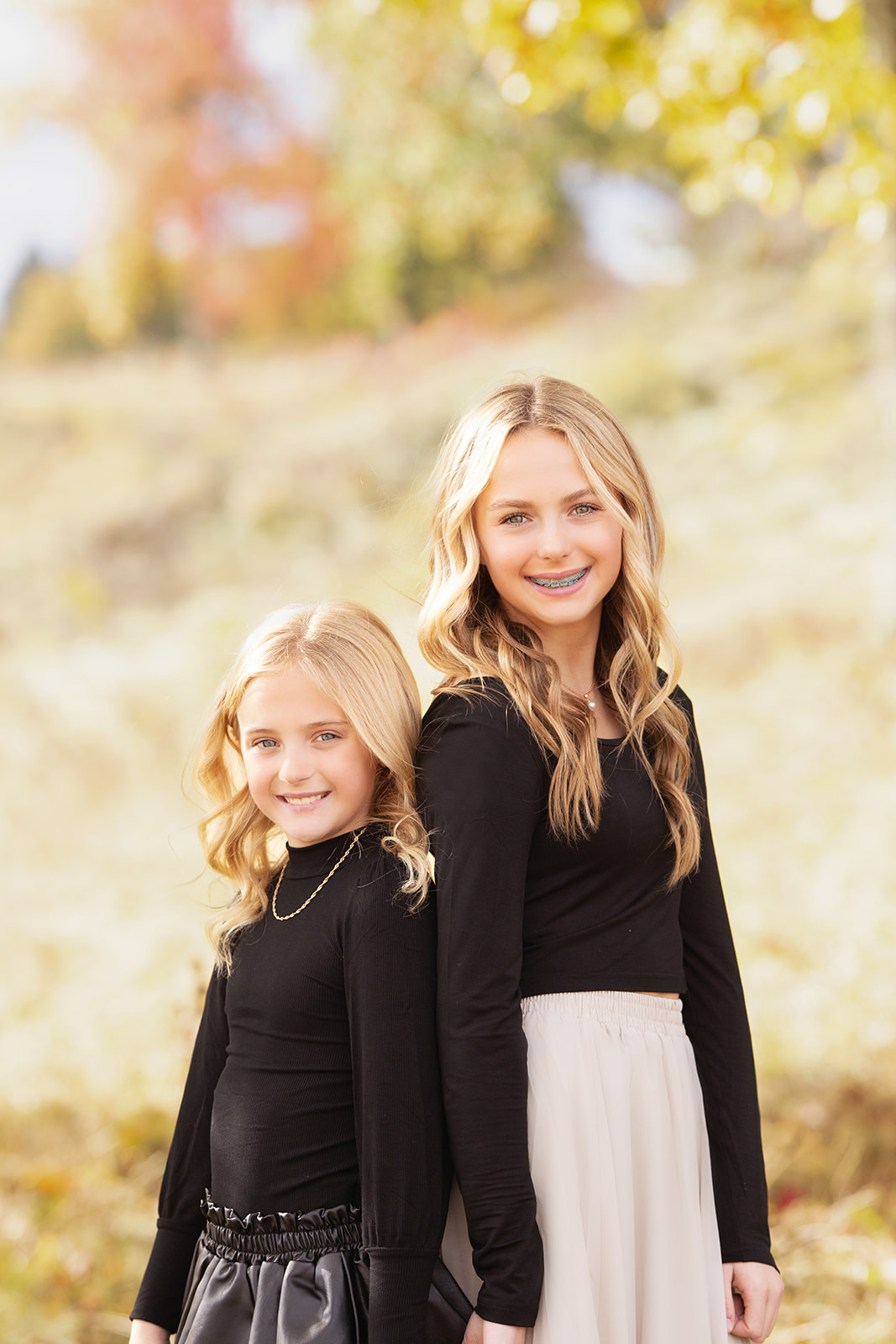 Smiling sisters in black tops stand back to back holding hands in a field at sunset after some Pittsburgh summer camps