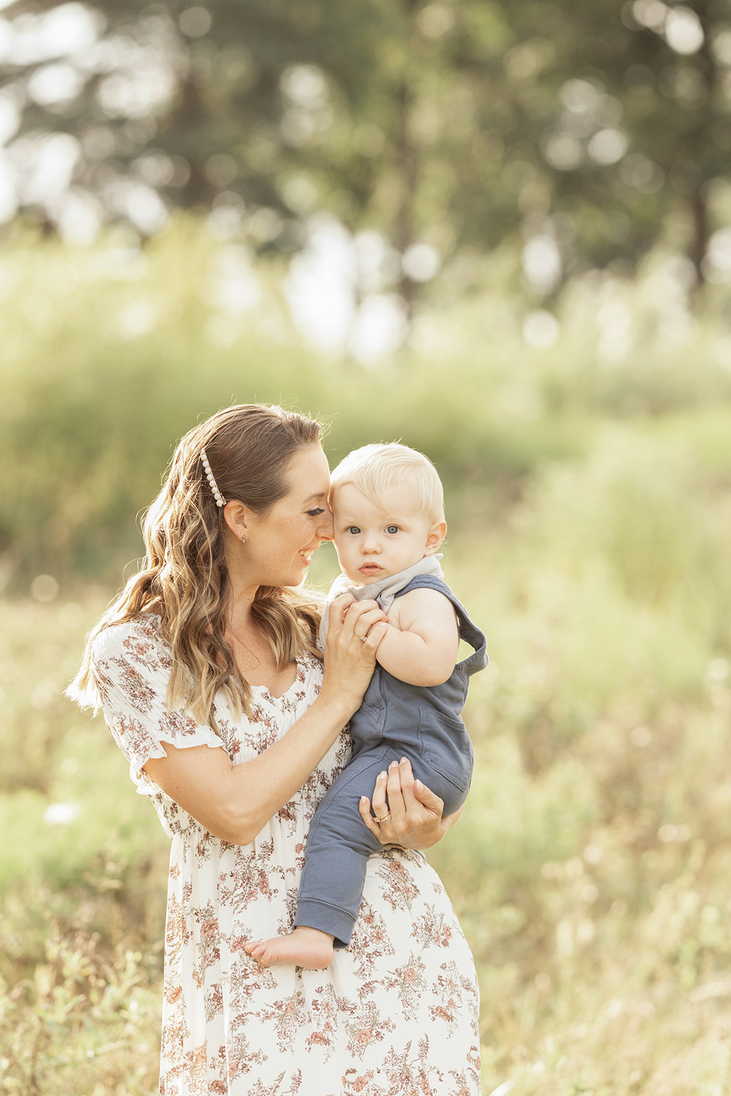 A happy mom snuggles her young toddler son on her hip at sunset in a field of tall grass