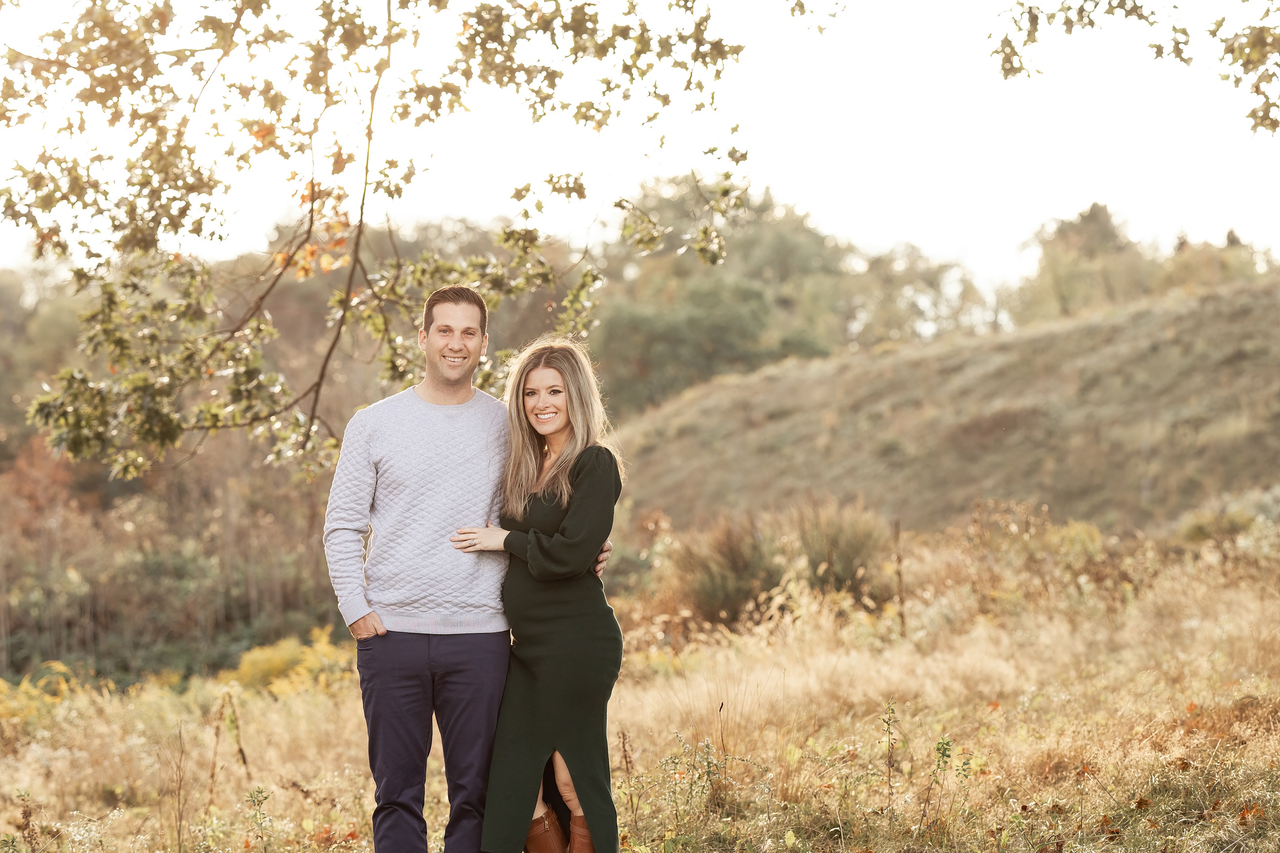 Happy expecting parents stand in a field of tall grasses on a hill at sunset holding one another