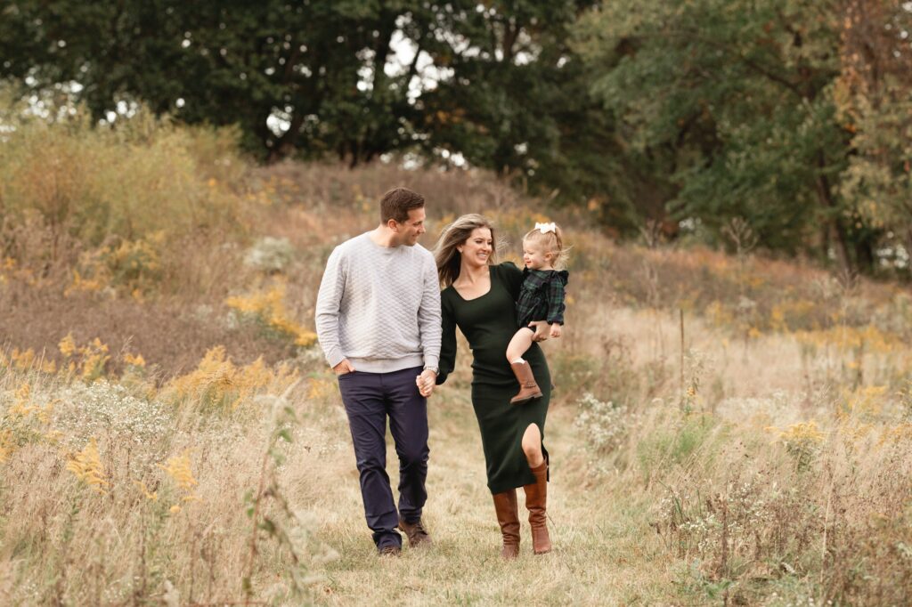 Happy parents walk in a park trail holding hands while giggling with their daughter on mom's hip after some toddler classes in Pittsburgh