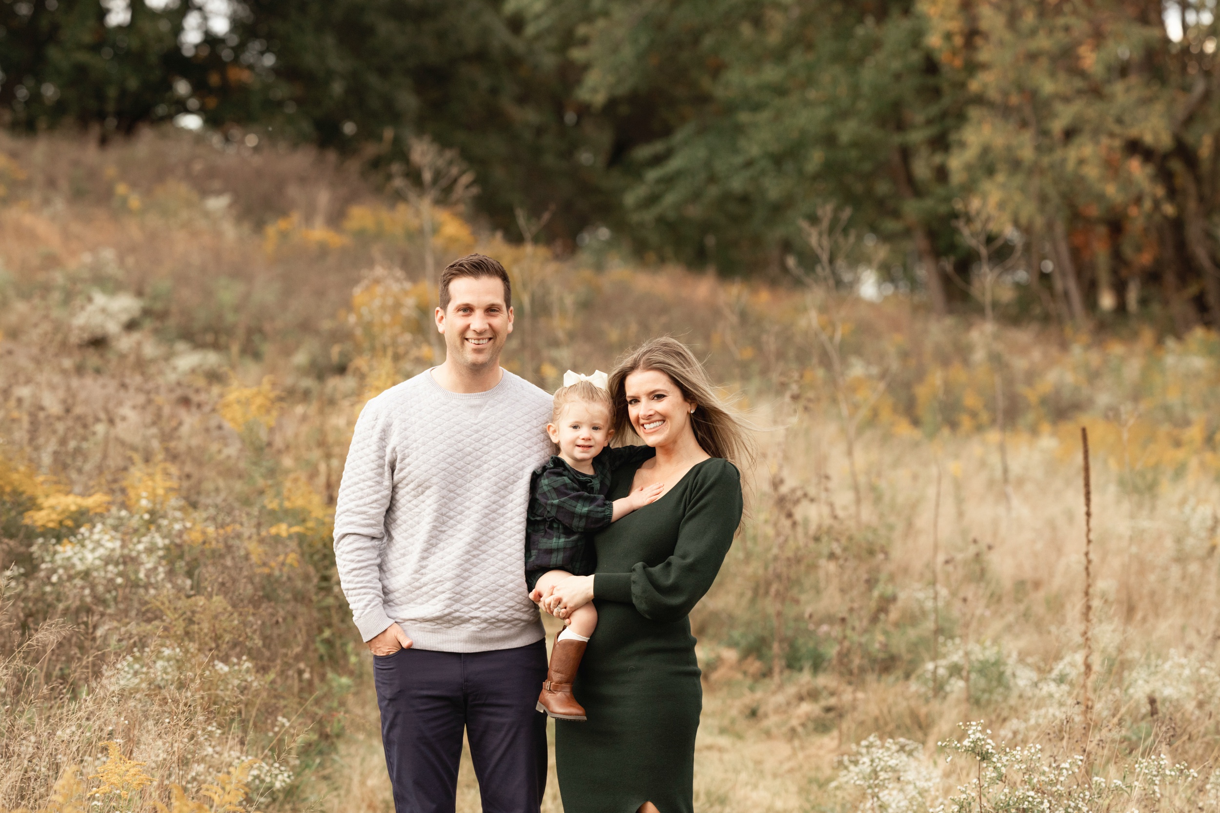 Happy mom and dad stand in a park trail with tall grasses holding their daughter in a plaid dress after some toddler classes in Pittsburgh