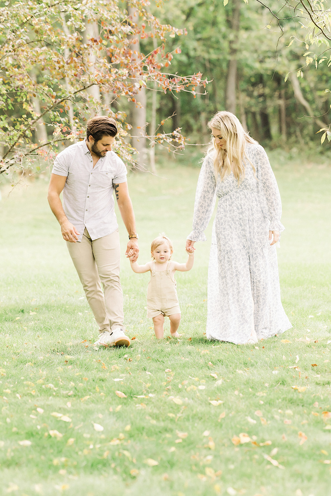 A happy toddler walks with mom and dad's help through a park lawn