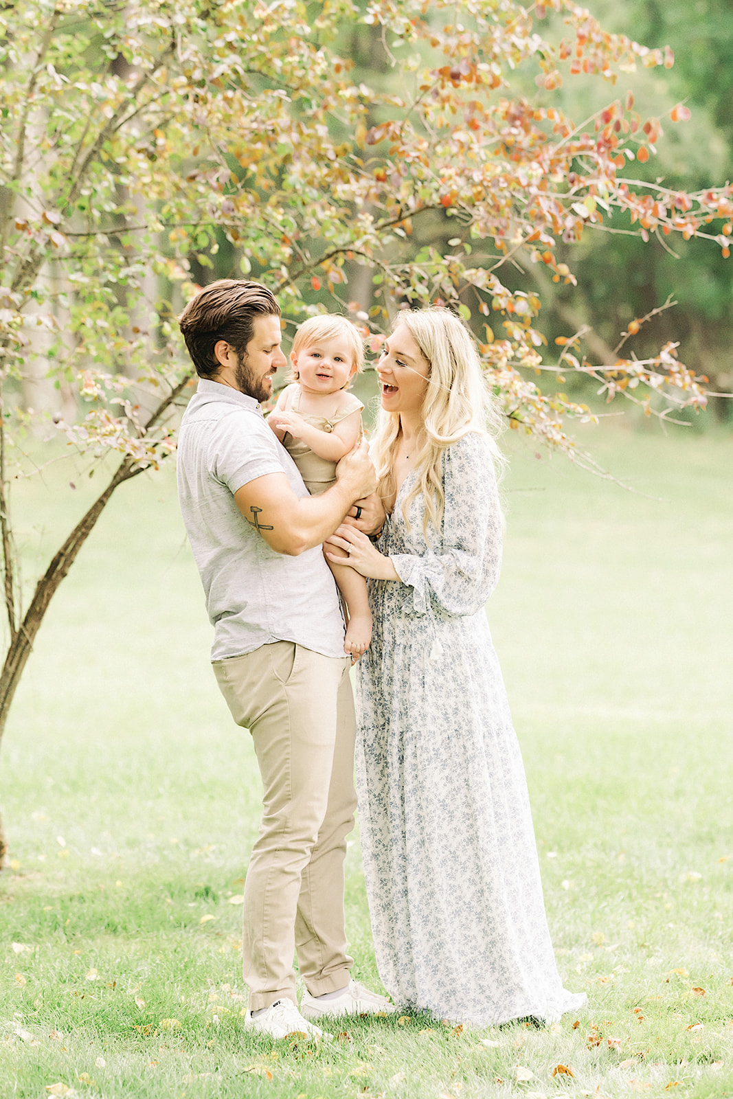 A happy mom and dad stand in a park under a young tree playing with their baby in dad's arms before visiting Pittsburgh trampoline parks