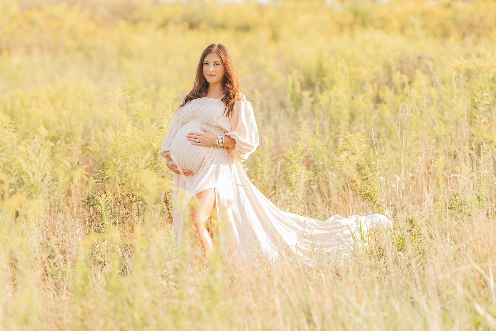 A pregnant woman walks in a field of tall grass in the sun with hands on her bump in a long flowing cream maternity gown