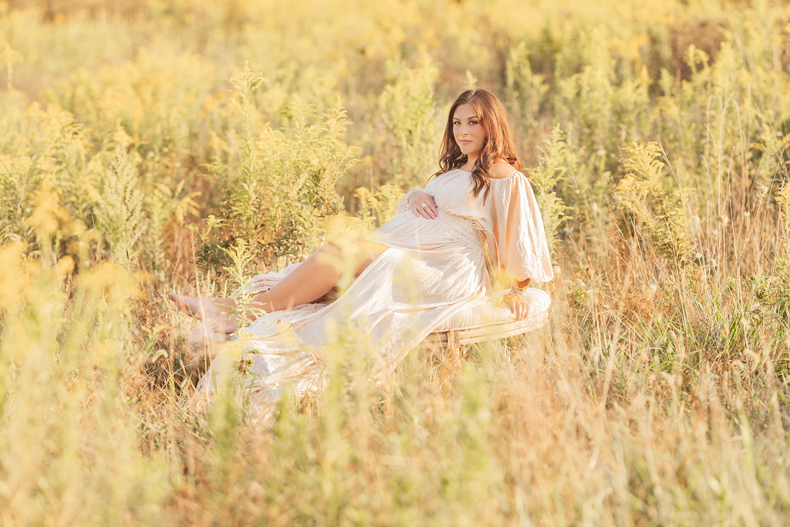 A mom to be sits across a bench in a field of tall grass in a cream maternity gown with a hand on her bump after visiting Pittsburgh yoga studios