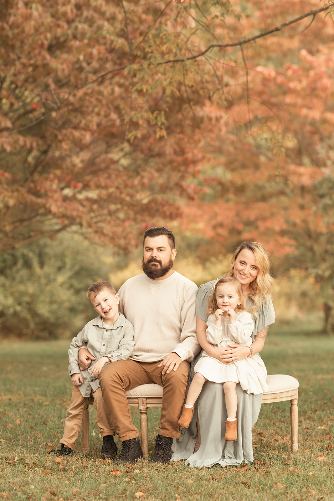 A happy family with 2 toddlers sits on a bench under a tree in a park smiling and giggling