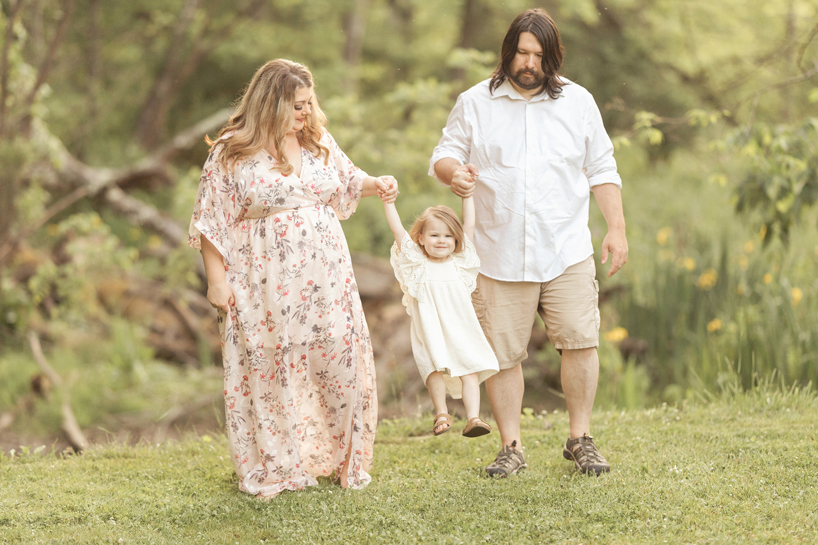 A toddler girl in a white dress swings between mom and dad by their hands