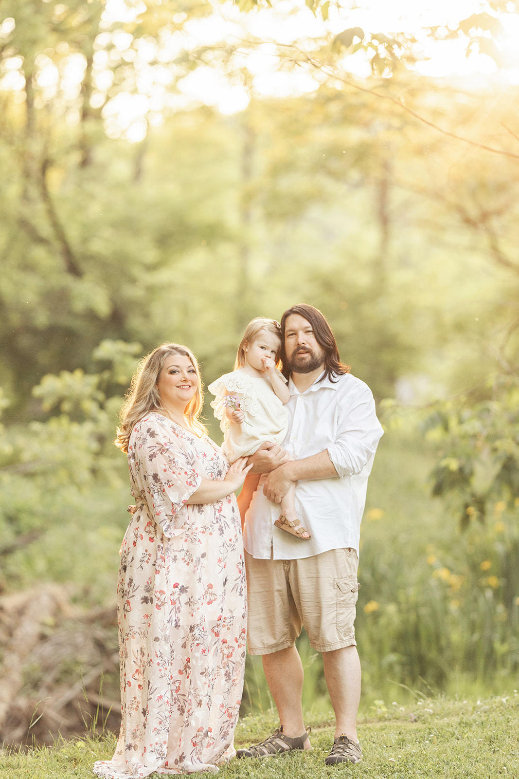 A happy mom in a floral dress stands with her husband holding their toddler daughter in a park at sunset during things to do in Pittsburgh with kids