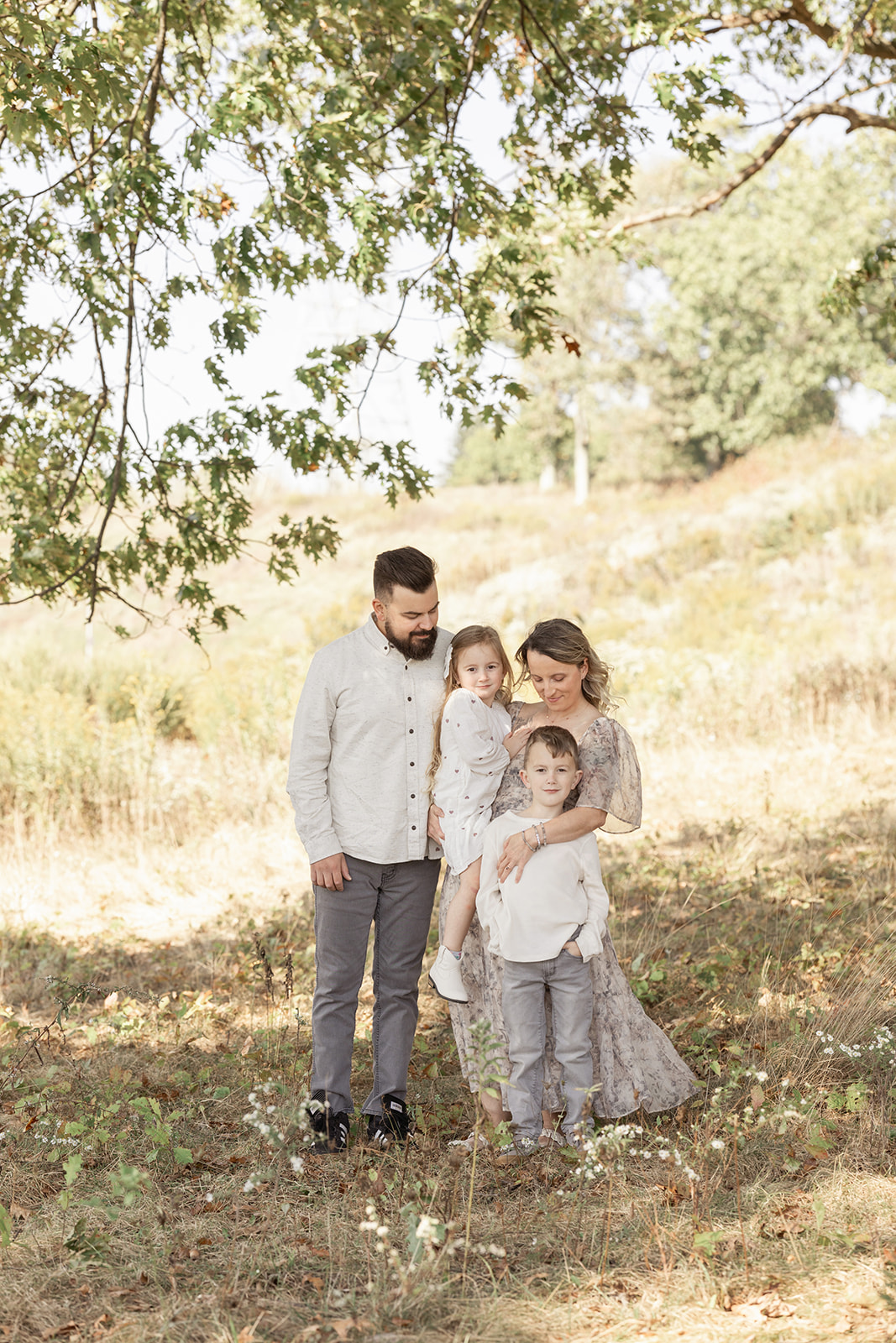 A happy family of 4 with two toddlers stand under a tree in a forest hugging and smiling