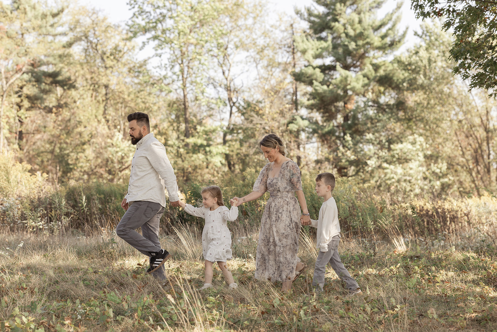 Happy mom and dad lead their toddler son and daughter through a forest at sunset holding hands before finding Christmas Lights in Pittsburgh