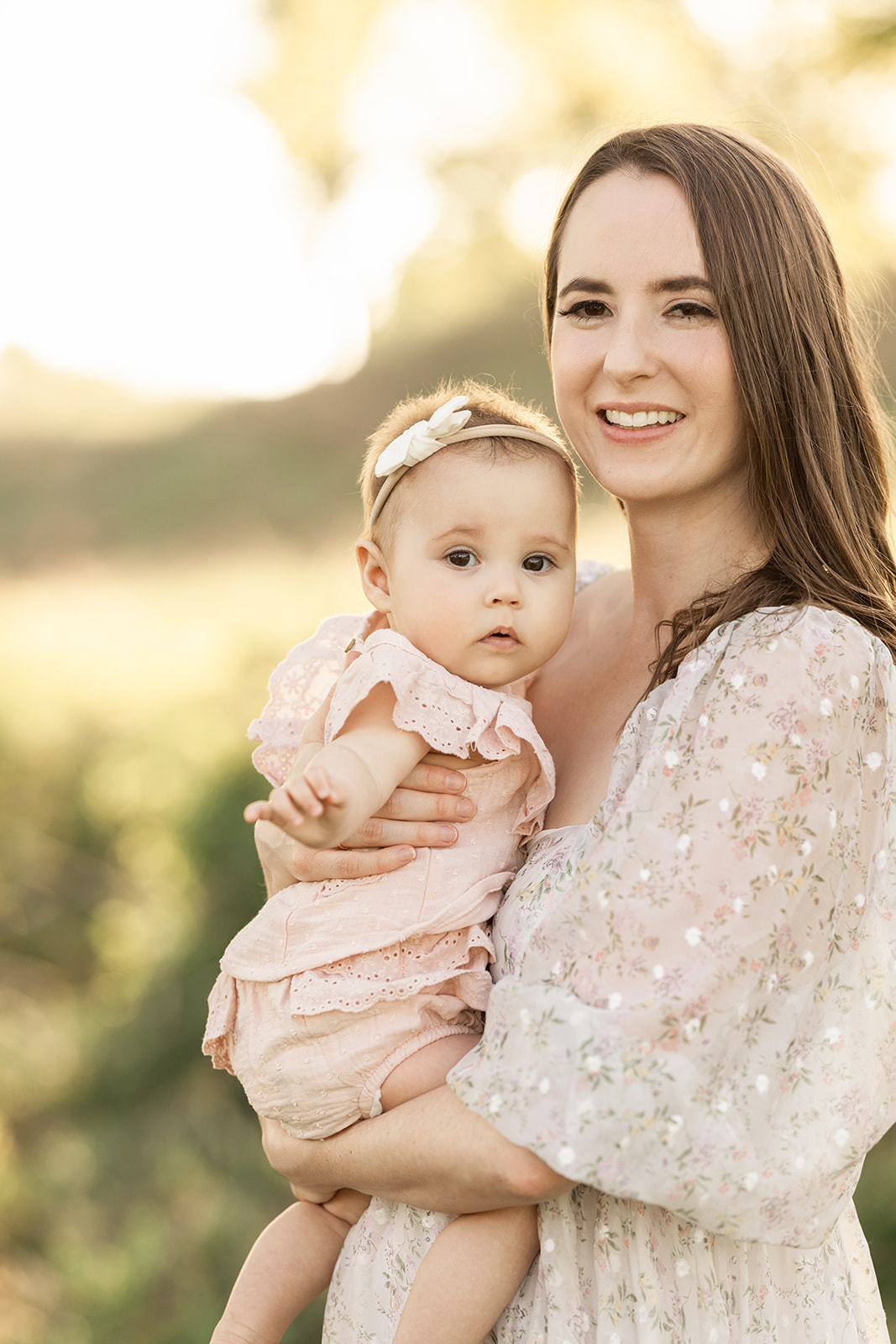 A smiling mom stands in a park at sunset holding her baby in a pink dress in her arms