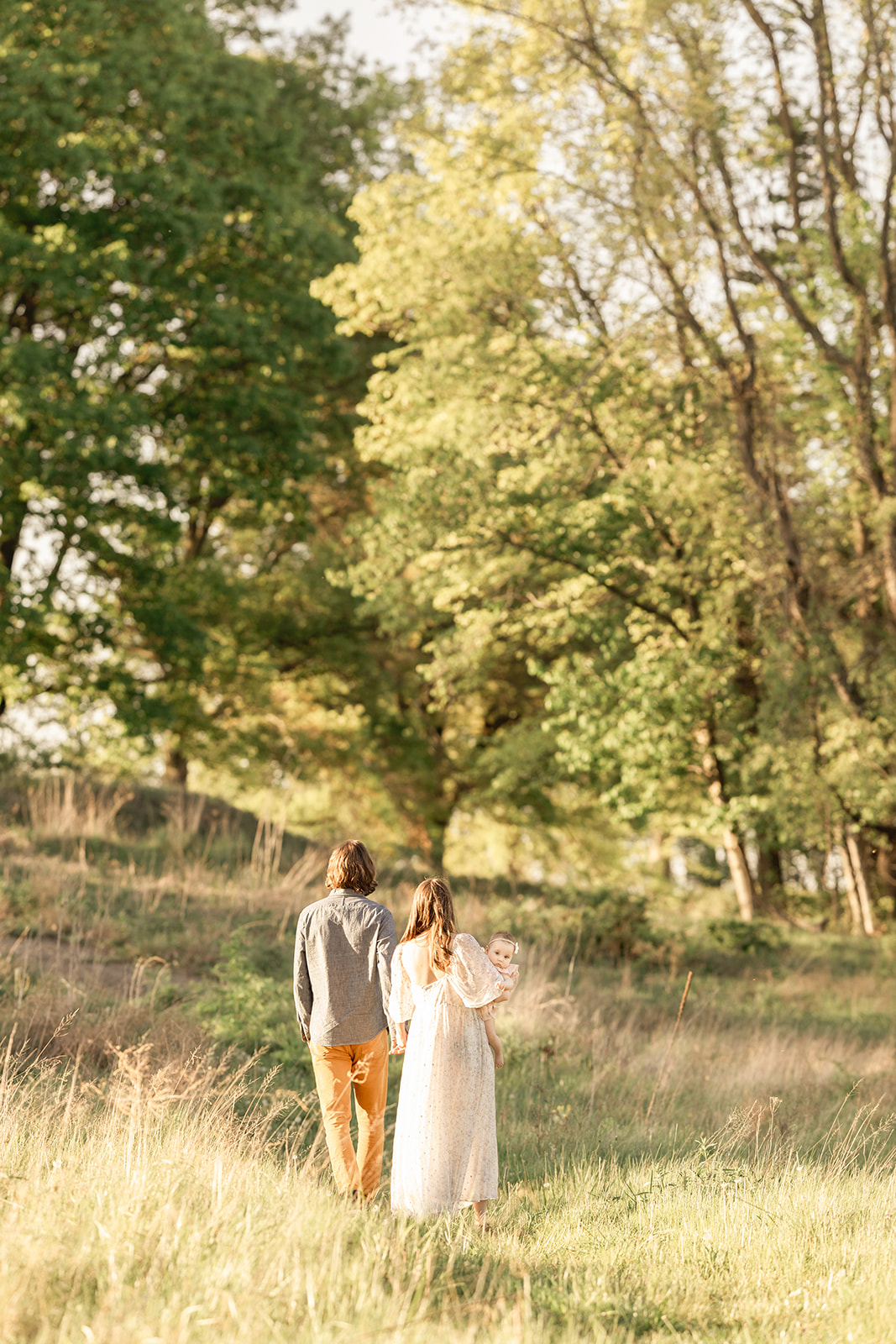 A mom and dad hold hands while walking in a forest at sunset with their baby on mom's hip before visiting Pittsburgh Christmas Tree Farms