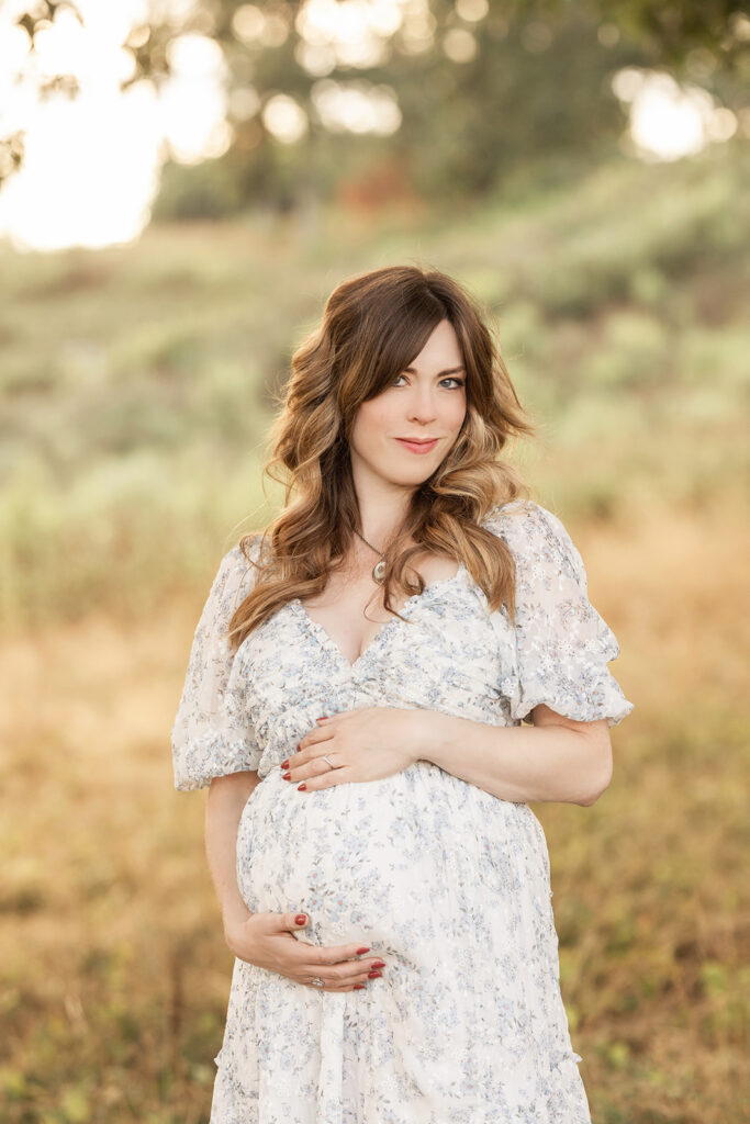 A mom to be in a floral print white maternity gown holds her bump and smiles under a tree at sunset after visiting All About Women Weirton, WV