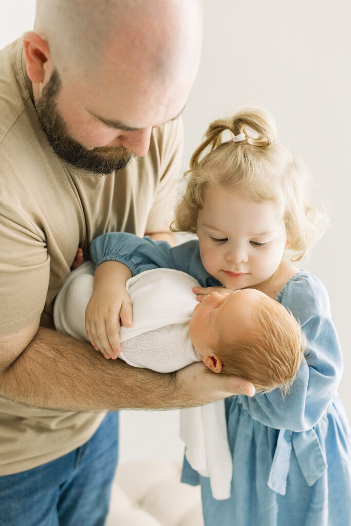 A young girl hugs her sleeping newborn baby sibling in dad's arms in a blue dress after visiting baby stores in Morgantown, WV