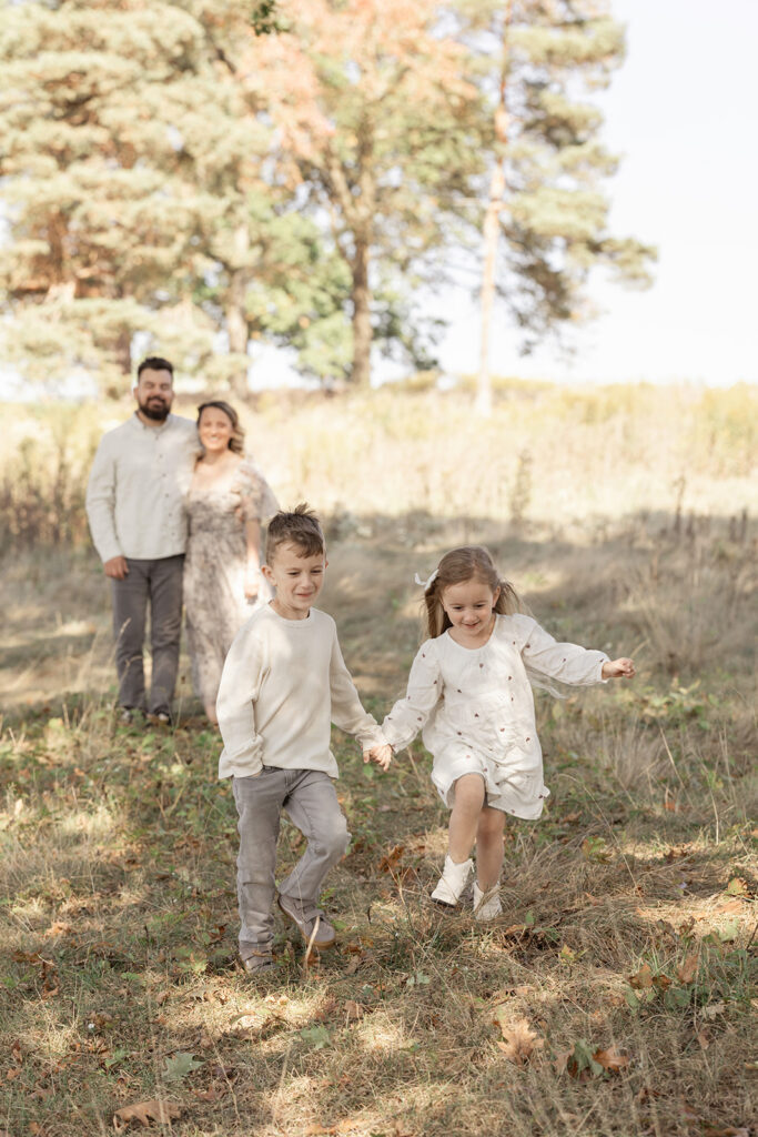 Toddler brother and sister run in a park holding hands away from happy mom and dad