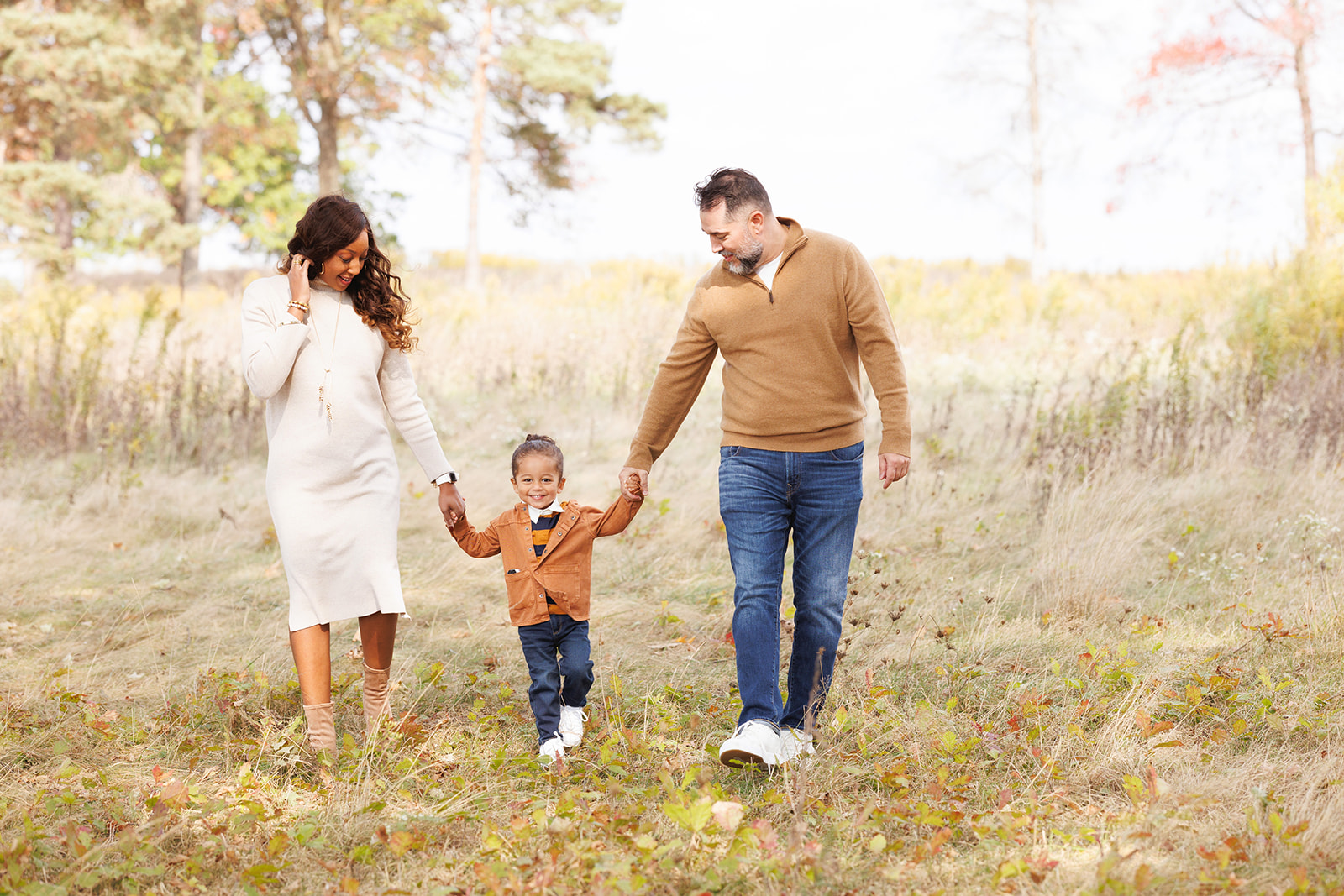 A happy toddler girl in blue pants and orange jacket walks in a park holding mom and dad's hands