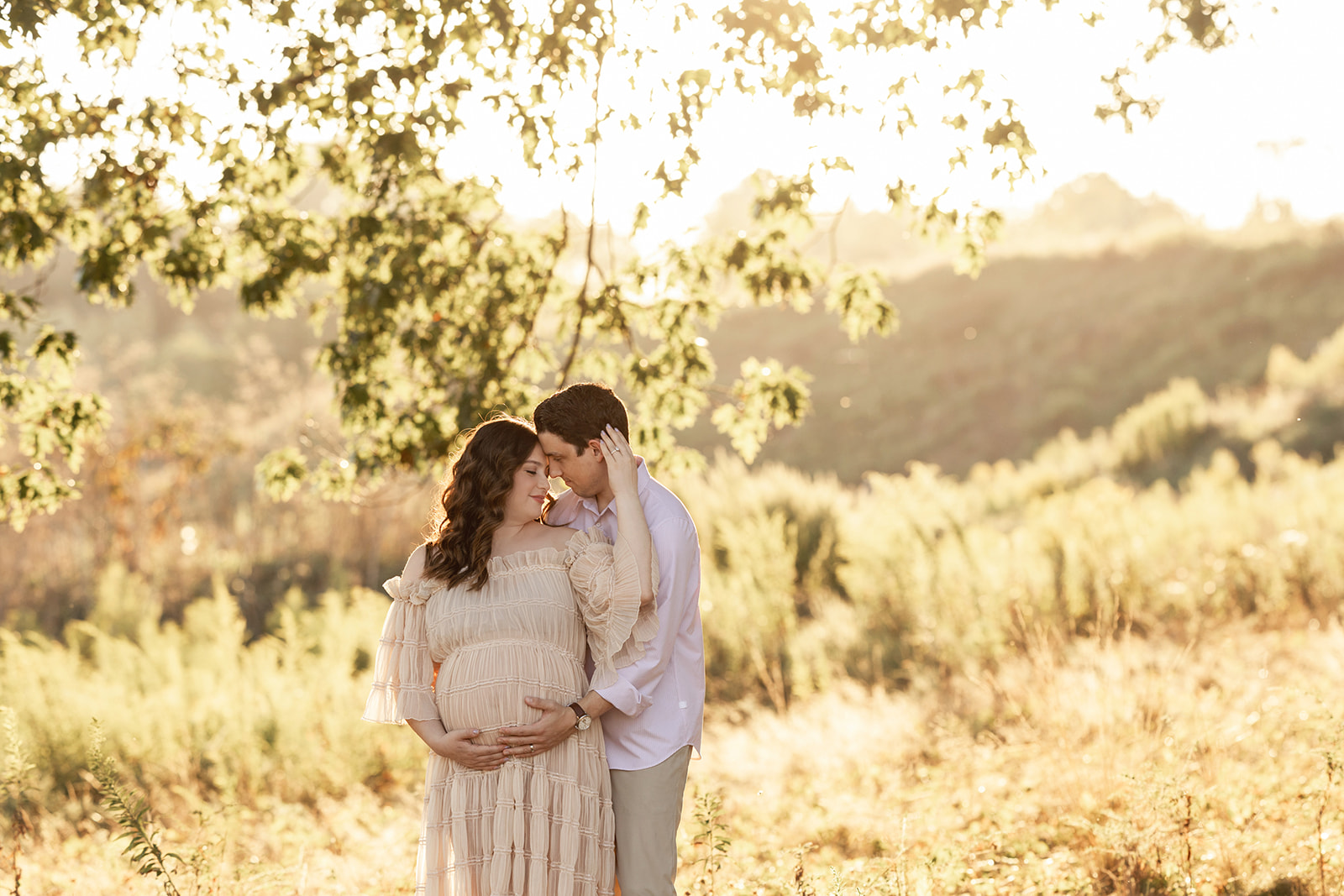 Expecting parents snuggle and hold the bump while standing under a tree at sunset on a hillside