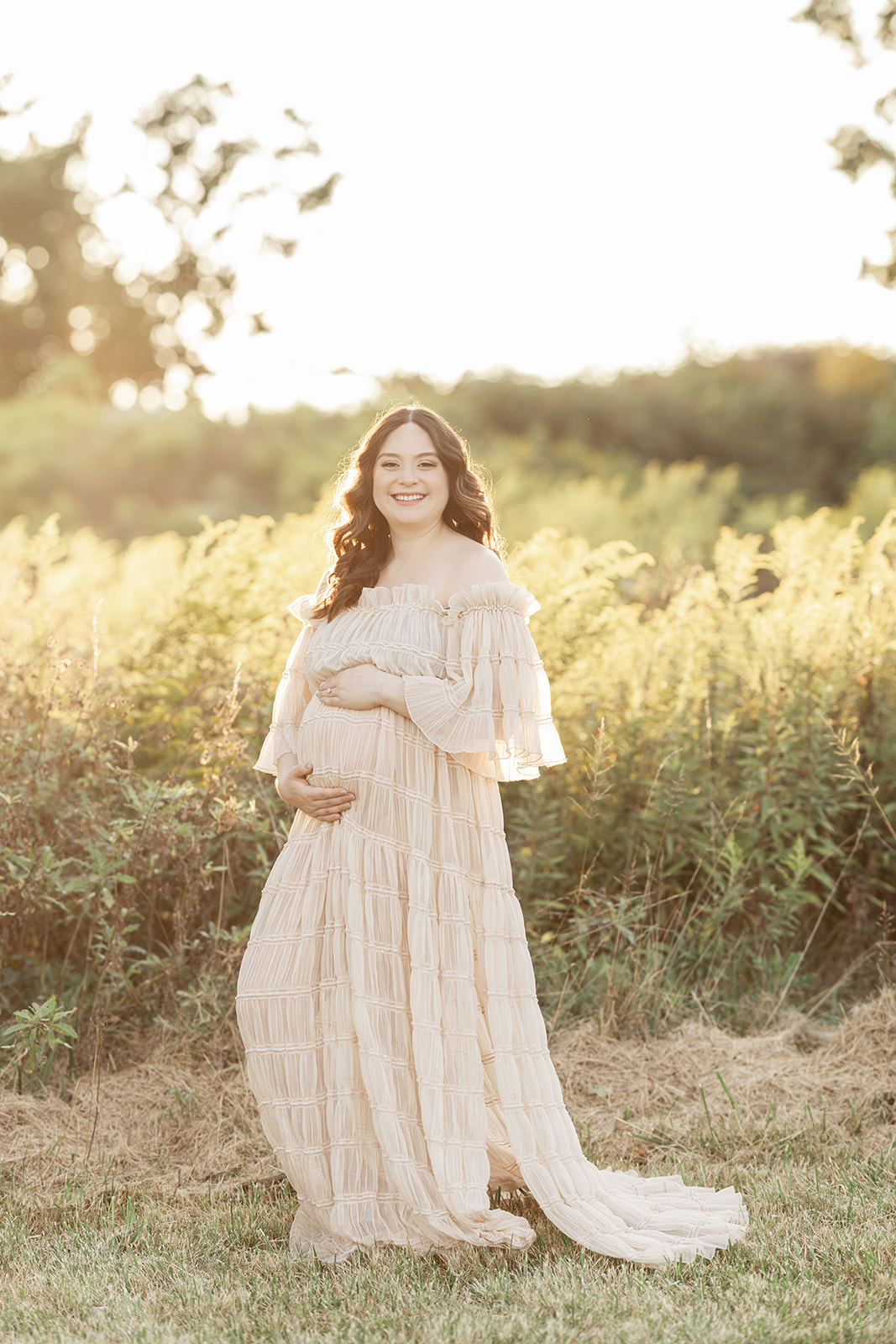 A smiling mother to be in a flowing maternity gown stands on the edge of tall grass at sunset thanks to a fertility clinic in Morgantown, WV