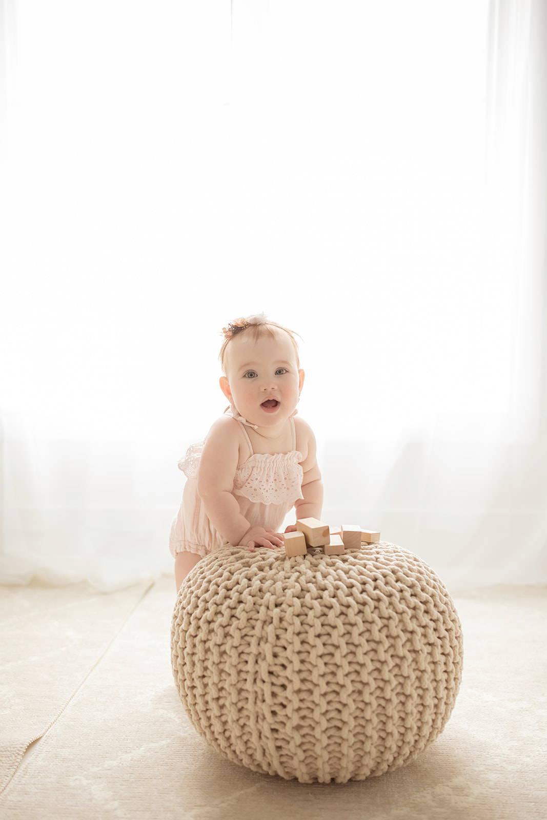 A baby girl stands with a woven stool while playing with wooden blocks in a studio under a window before visiting indoor playgrounds in Morgantown, WV