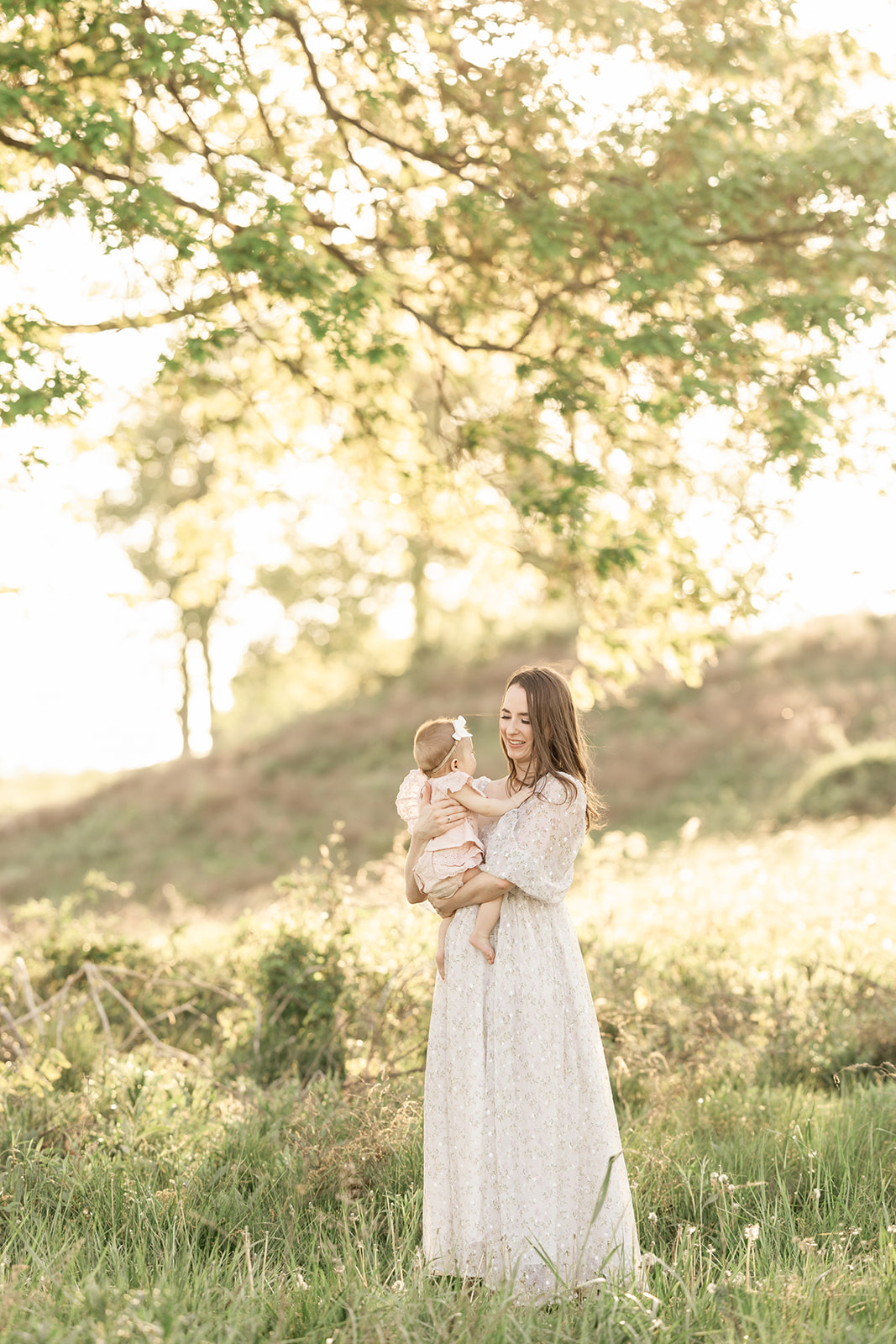 A woman in a lace dress holds her infant daughter in a pink onesie under a tree at sunset during kid-friendly things to do in Morgantown, WV