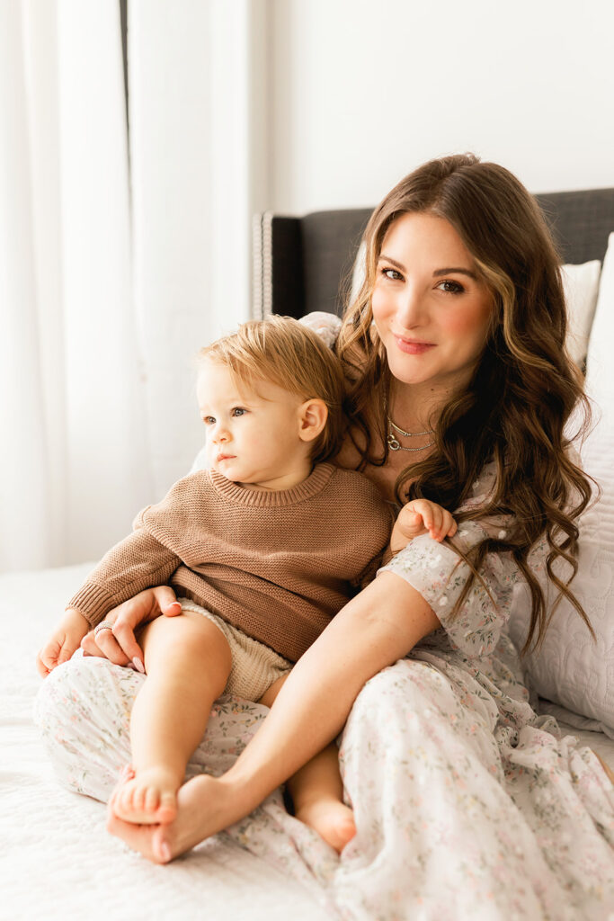 A smiling mother cuddles her infant in her lap in a flowing dress and brown sweater