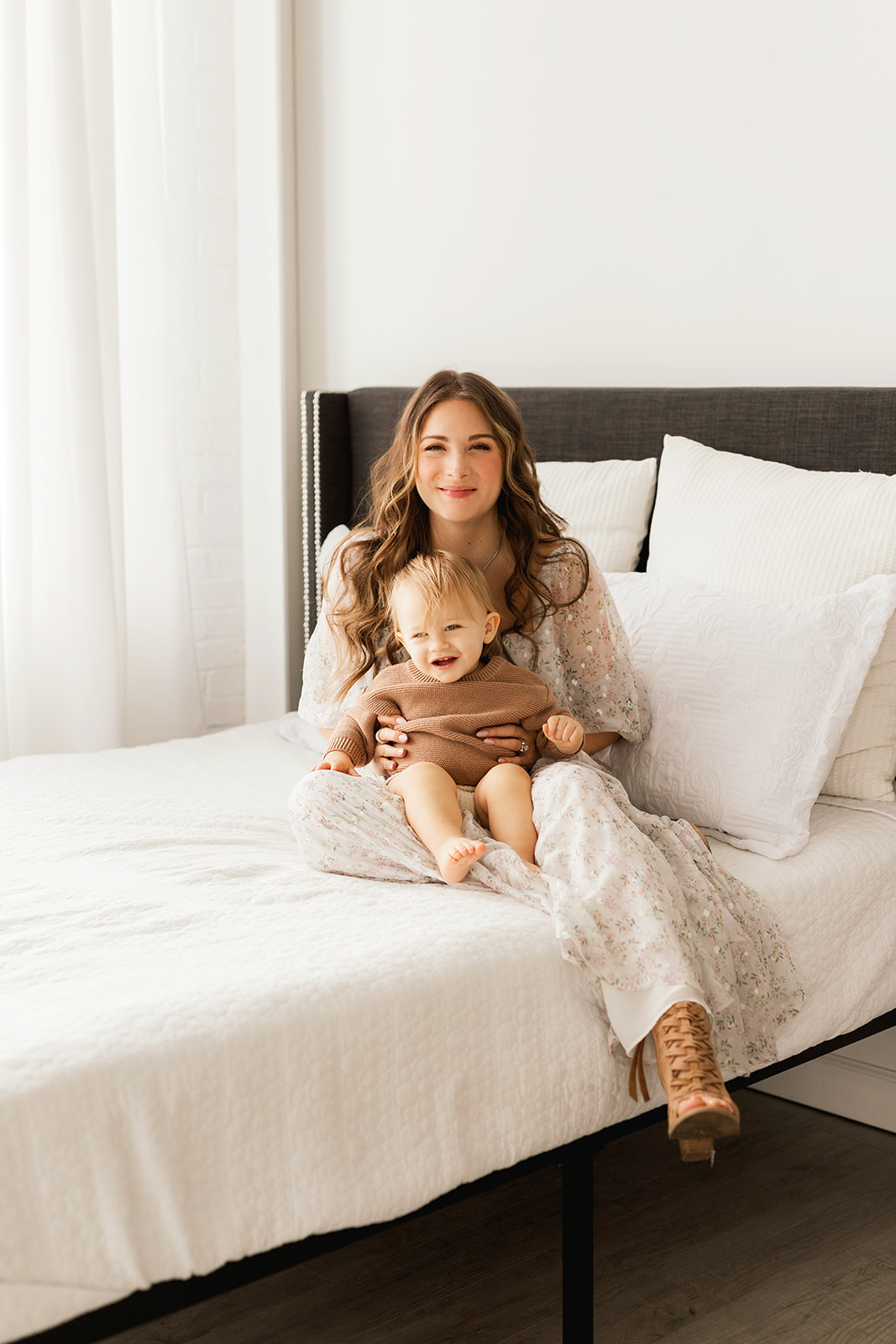 A smiling mother sits with her happy infant in her lap on a bed in a long dress after finding Morgantown, WV pediatricians