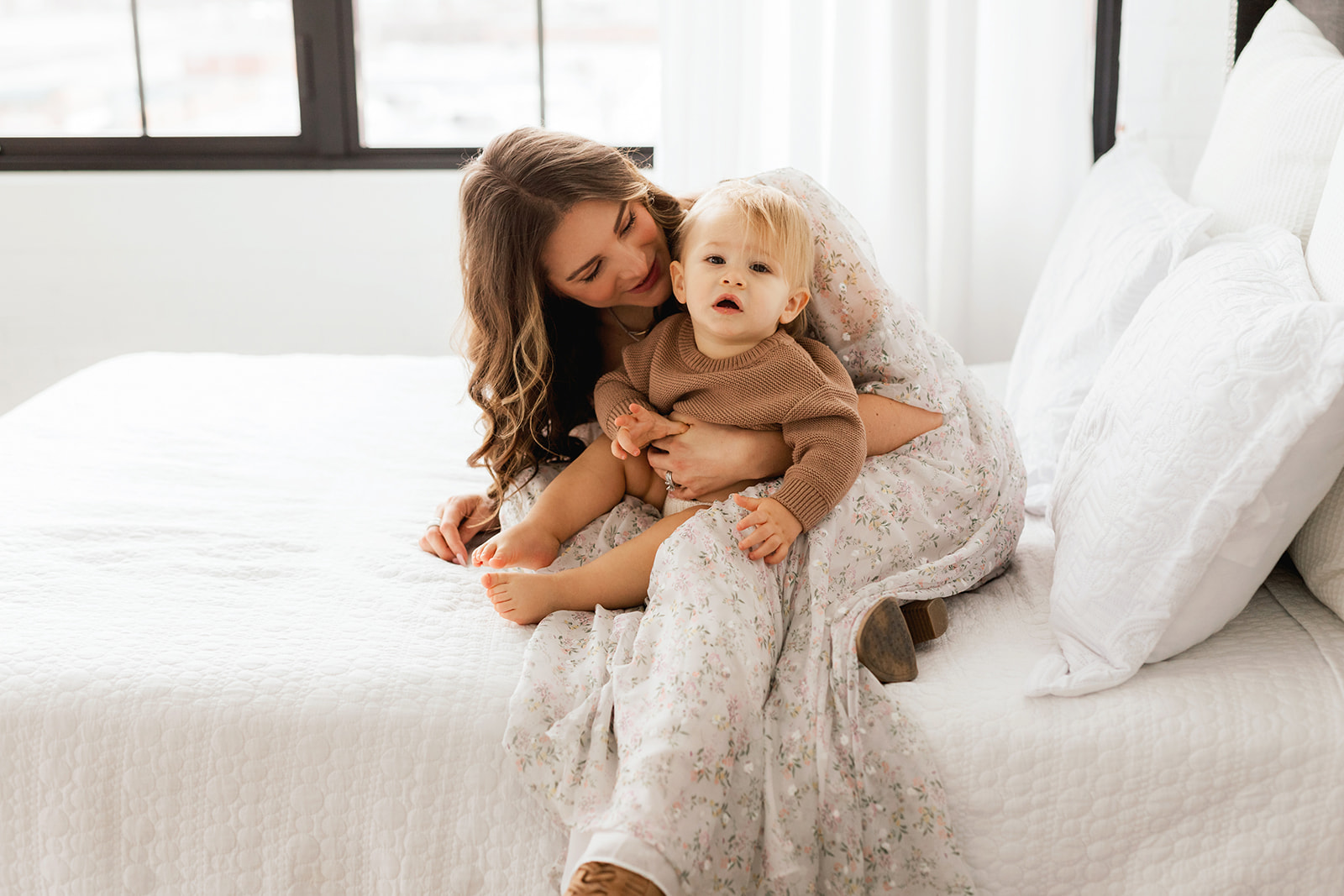 A happy mom plays with her toddler in a brown sweater on a white bed after visiting Morgantown, WV pediatricians