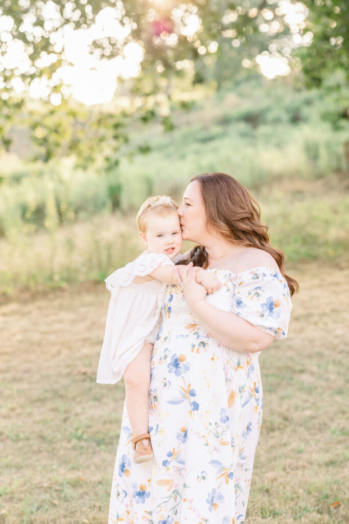 mom kissing daughter's head in field at sunset with petite magnolia photography