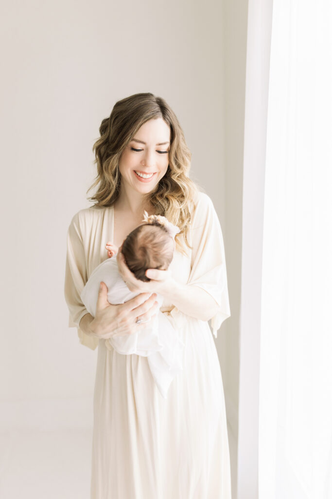 mom in cream flowy gown holding sewickley newborn baby girl swaddled in cream wrap by natural window light at petite magnolia photography